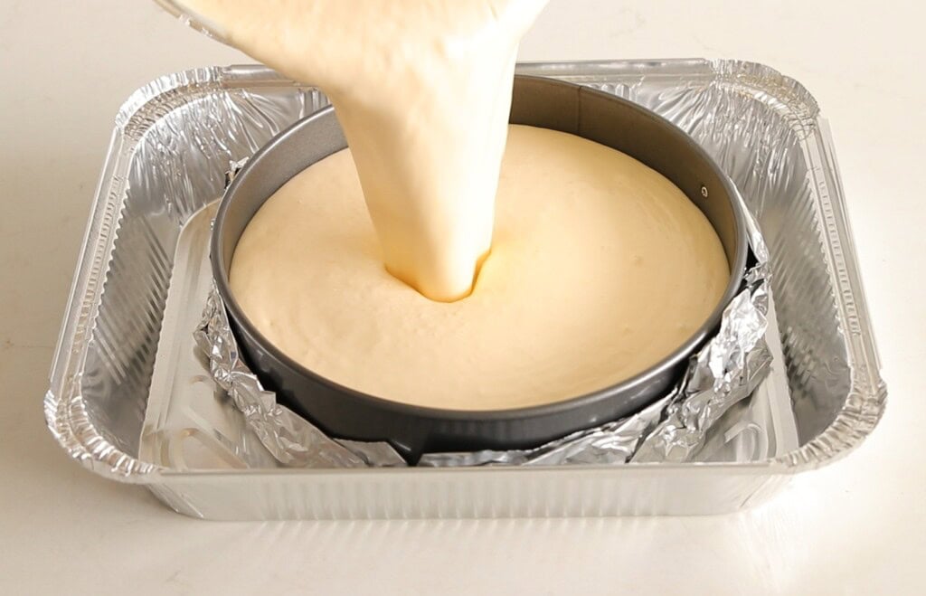 Batter is being poured into a springform pan lined with foil, which sits in a larger aluminum tray, likely for a water bath, on a white surface.