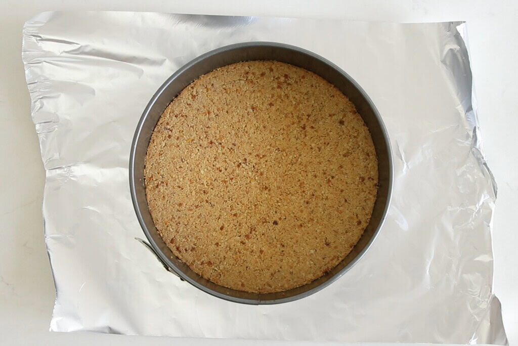 A round springform pan with a pressed crumb crust sits on a sheet of aluminum foil, viewed from above on a white surface.