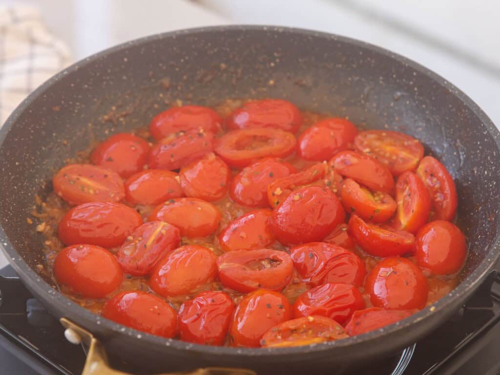 A black frying pan filled with halved cherry tomatoes cooking in a sauce on a stovetop, with some herbs and seasonings visible.
