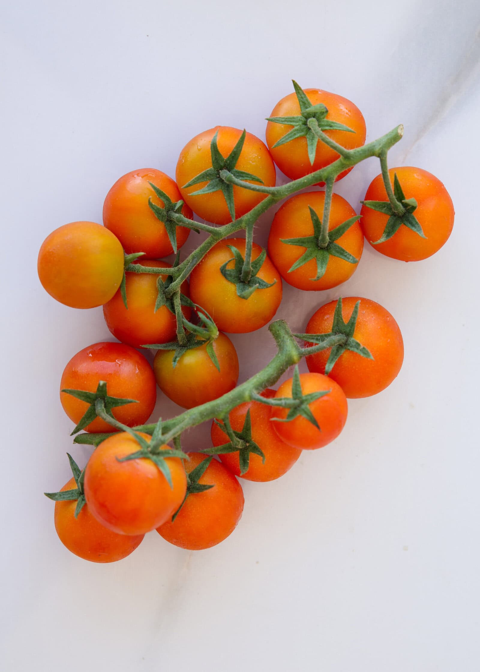 A cluster of orange-red cherry tomatoes still attached to green stems, arranged on a white marble surface.