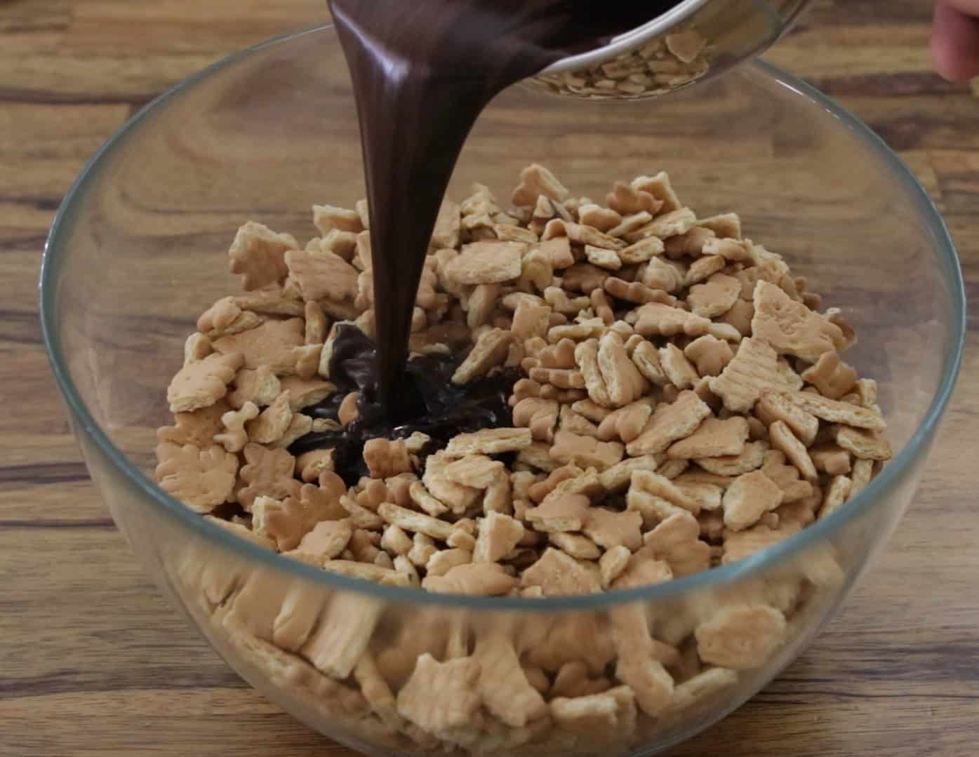 A clear glass bowl filled with broken graham crackers has chocolate sauce being poured over them, set on a wooden surface.