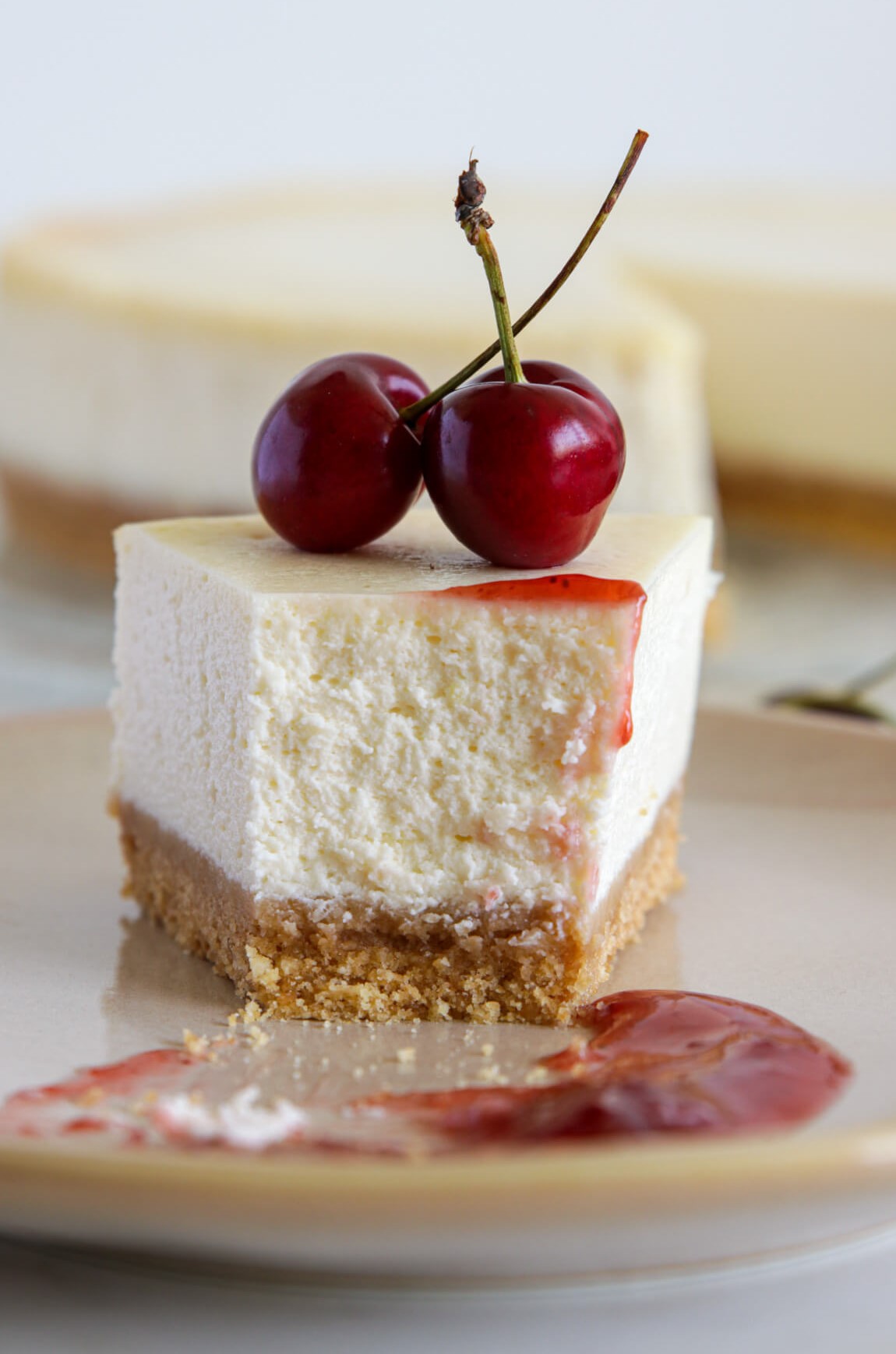 A slice of creamy cheesecake with a graham cracker crust is topped with two fresh cherries and cherry sauce, sitting on a plate with a smear of sauce beside it. The rest of the cheesecake is blurred in the background.