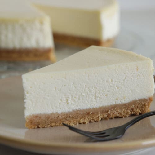 A slice of classic cheesecake with a thick, creamy layer sits on a beige plate next to a fork. The cheesecake has a golden brown crust and another slice is visible in the background.
