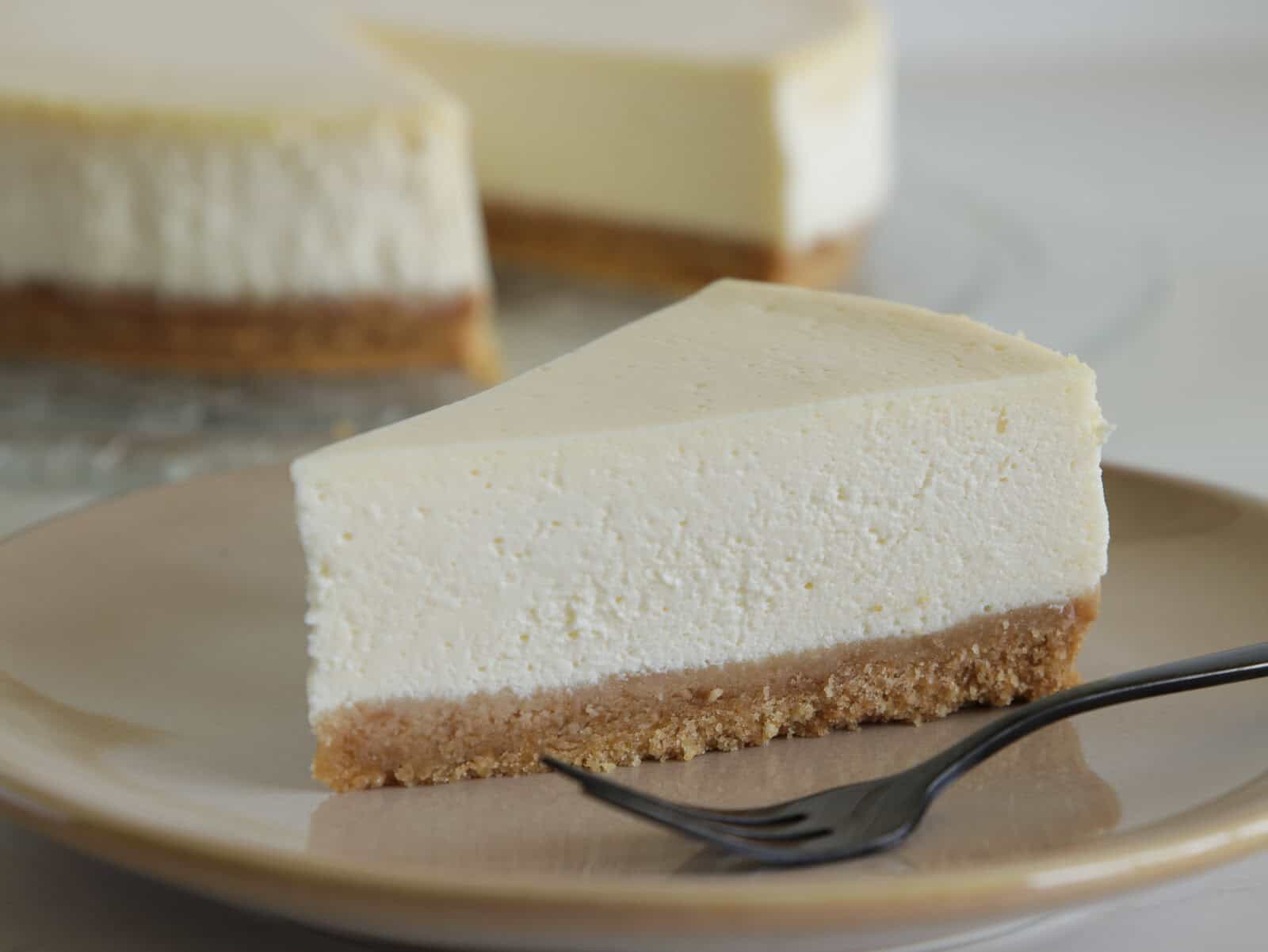 A slice of classic cheesecake with a thick, creamy layer sits on a beige plate next to a fork. The cheesecake has a golden brown crust and another slice is visible in the background.
