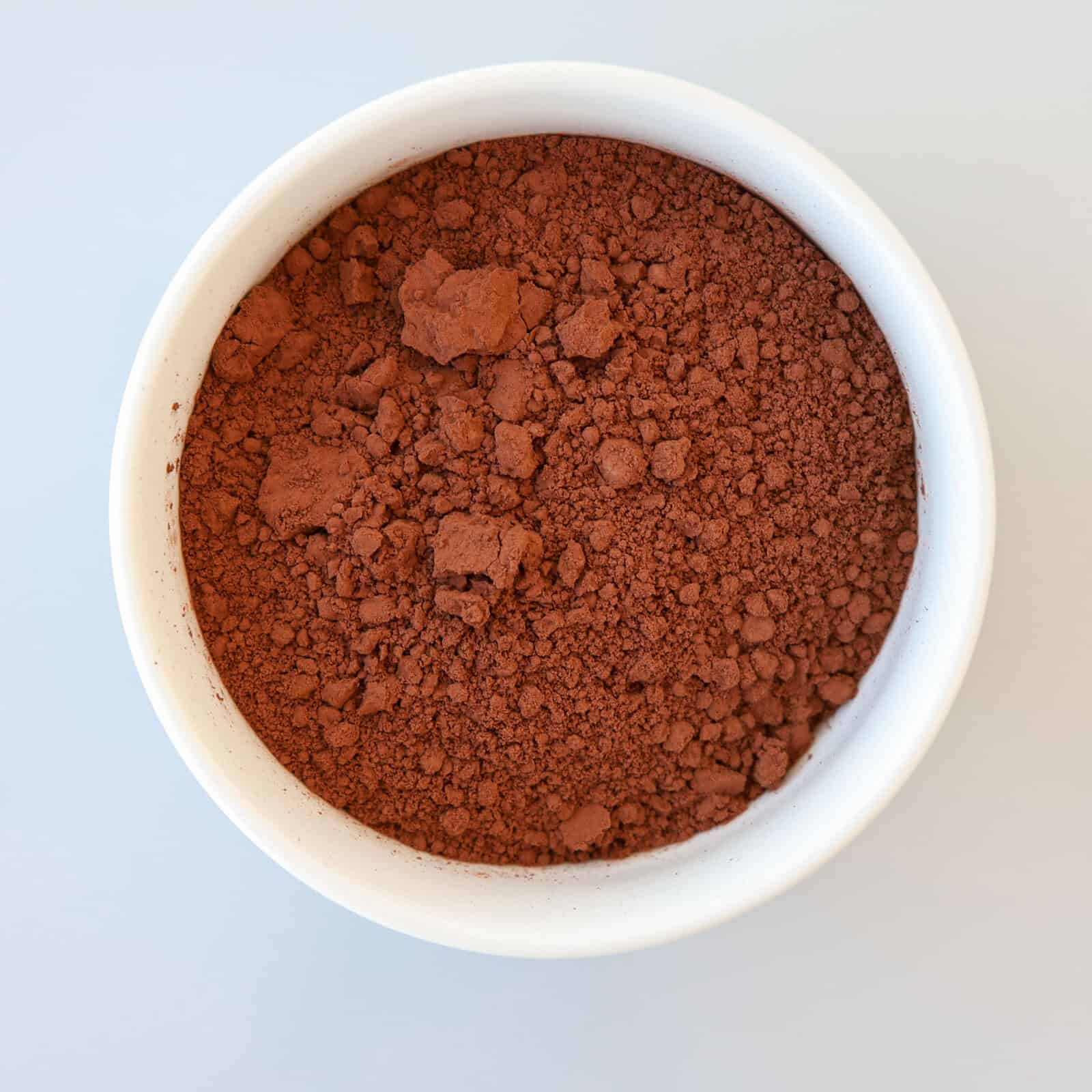 A white bowl filled with loose, brown cocoa powder seen from above on a light background.