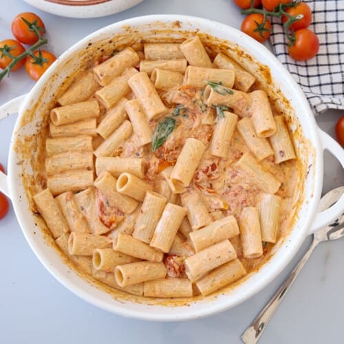 A white dish filled with creamy rigatoni pasta in a tomato sauce, garnished with basil. Cherry tomatoes and a checkered cloth are nearby, with a spoon resting beside the dish.