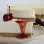 A slice of cheesecake with a graham cracker crust topped with two cherries and cherry sauce, served on a plate. The rest of the cheesecake and some cherries are visible in the background.