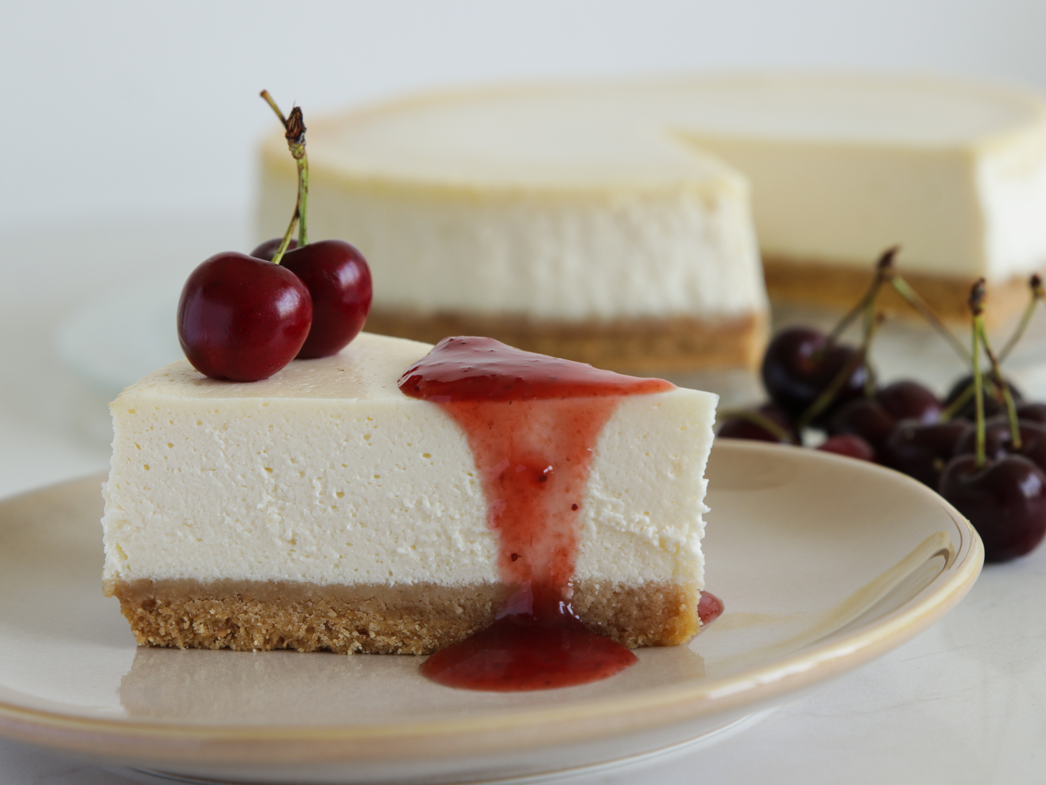 A slice of cheesecake with a graham cracker crust topped with two cherries and cherry sauce, served on a plate. The rest of the cheesecake and some cherries are visible in the background.