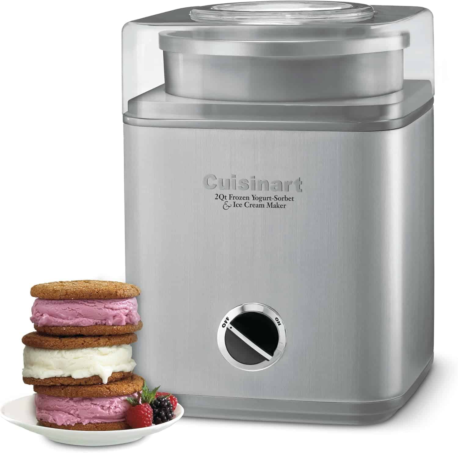 A stainless steel Cuisinart ice cream maker with a clear lid is shown next to three ice cream cookie sandwiches and a few fresh berries. The control knob is set to the "on" position.
