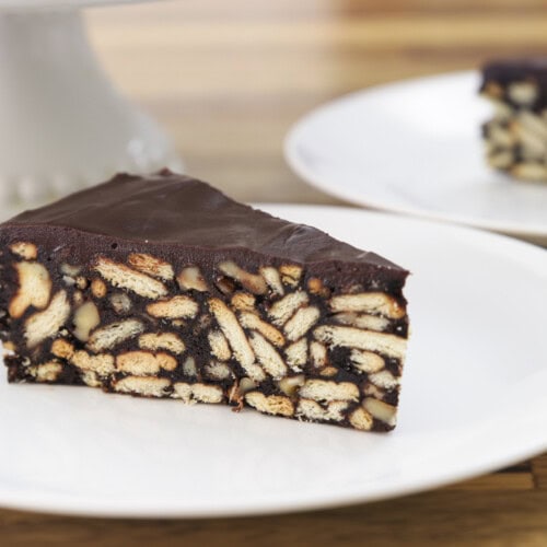 A slice of chocolate biscuit cake (lazy cake) with visible biscuit pieces and nuts sits on a white plate, with another plate of cake blurred in the background.