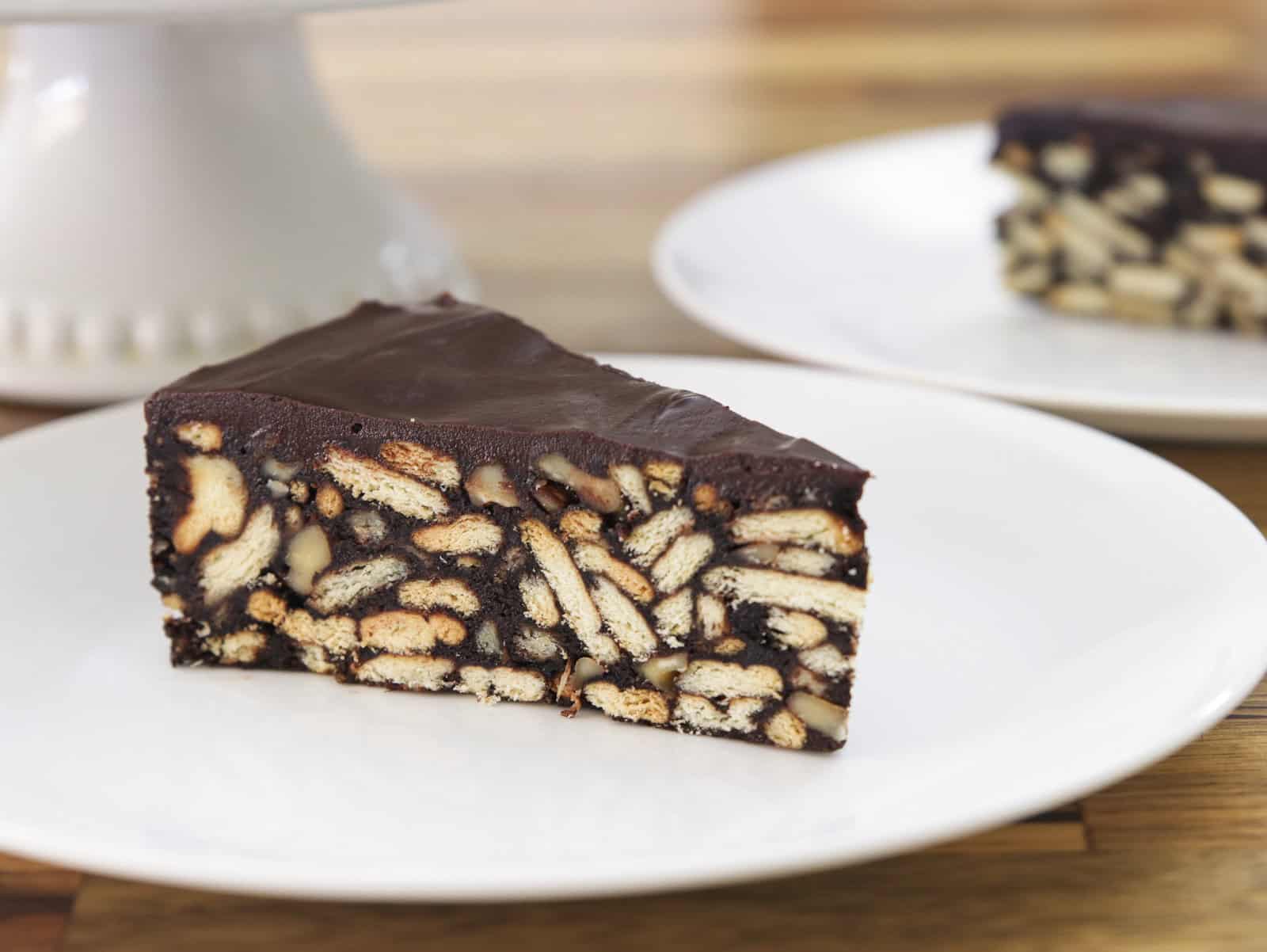 A slice of chocolate biscuit cake (lazy cake) with visible biscuit pieces and nuts sits on a white plate, with another plate of cake blurred in the background.