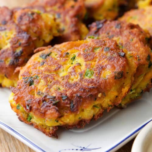 Golden brown vegetable fritters with flecks of green herbs are stacked on a white plate with a blue trim, showing a crispy exterior and slightly uneven, rustic shapes.