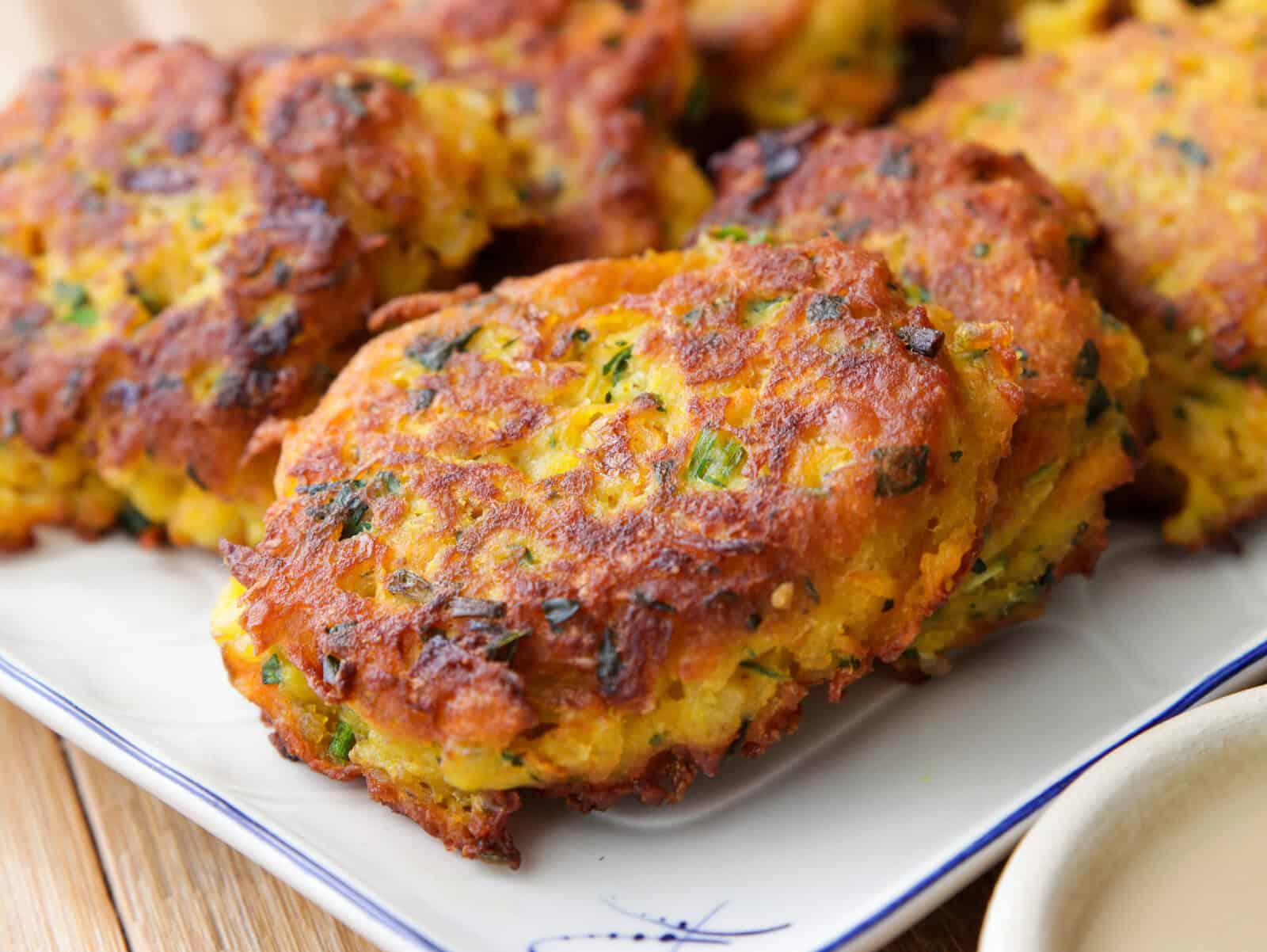 Golden brown baked lentil fritters with flecks of green herbs are stacked on a white plate with a blue trim, showing a crispy exterior and slightly uneven, rustic shapes.