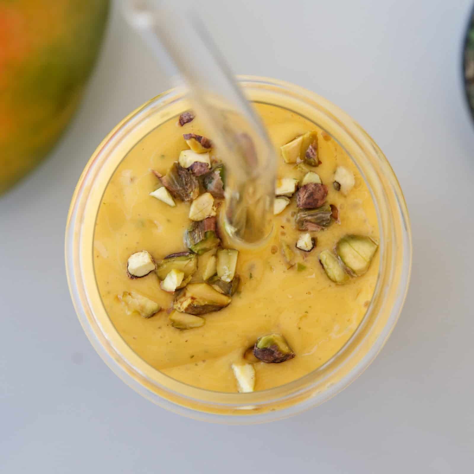 A top view of a glass filled with creamy yellow mango lassi, topped with chopped pistachios and a glass straw, with part of a mango visible in the corner.