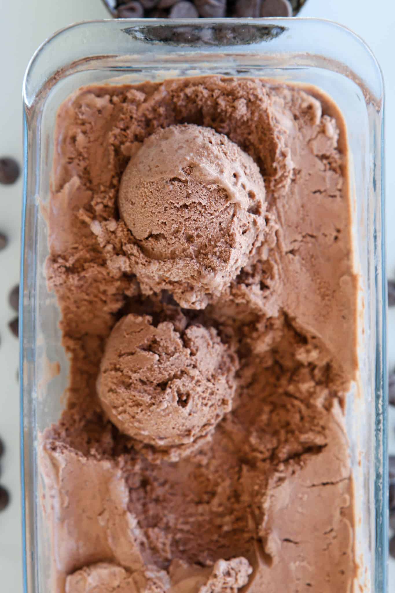 Two scoops of chocolate ice cream in a glass container, with chocolate chips scattered around on a white surface.