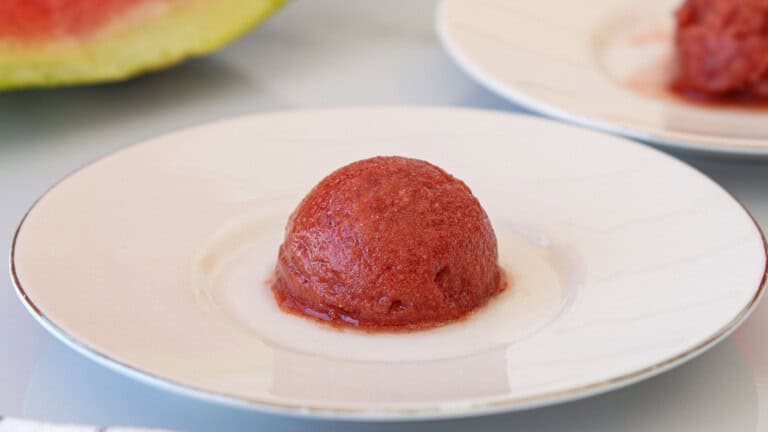 A scoop of watermelon sorbet sits on a white plate, with part of a watermelon and another scoop visible in the background.