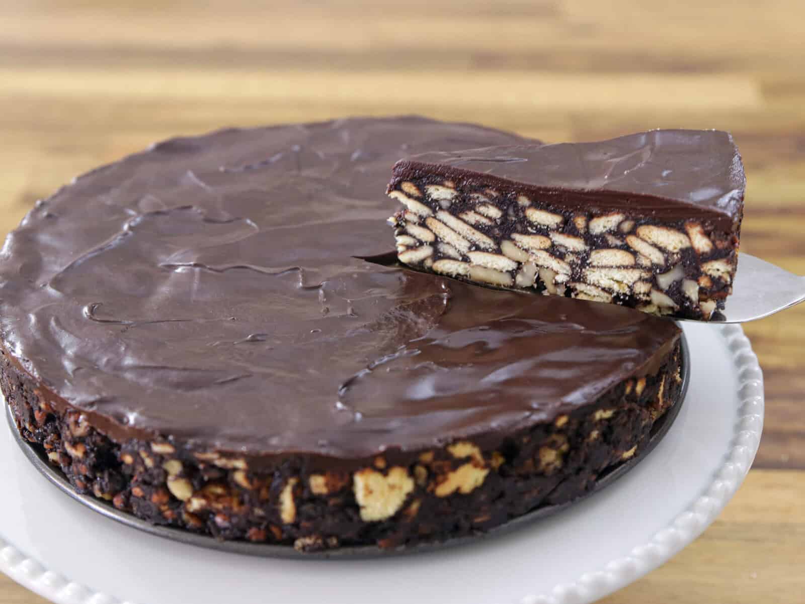 A no-bake chocolate biscuit cake with a glossy chocolate coating, featuring visible pieces of biscuit inside. One slice is being lifted to show the interior texture. The cake sits on a white plate on a wooden surface.