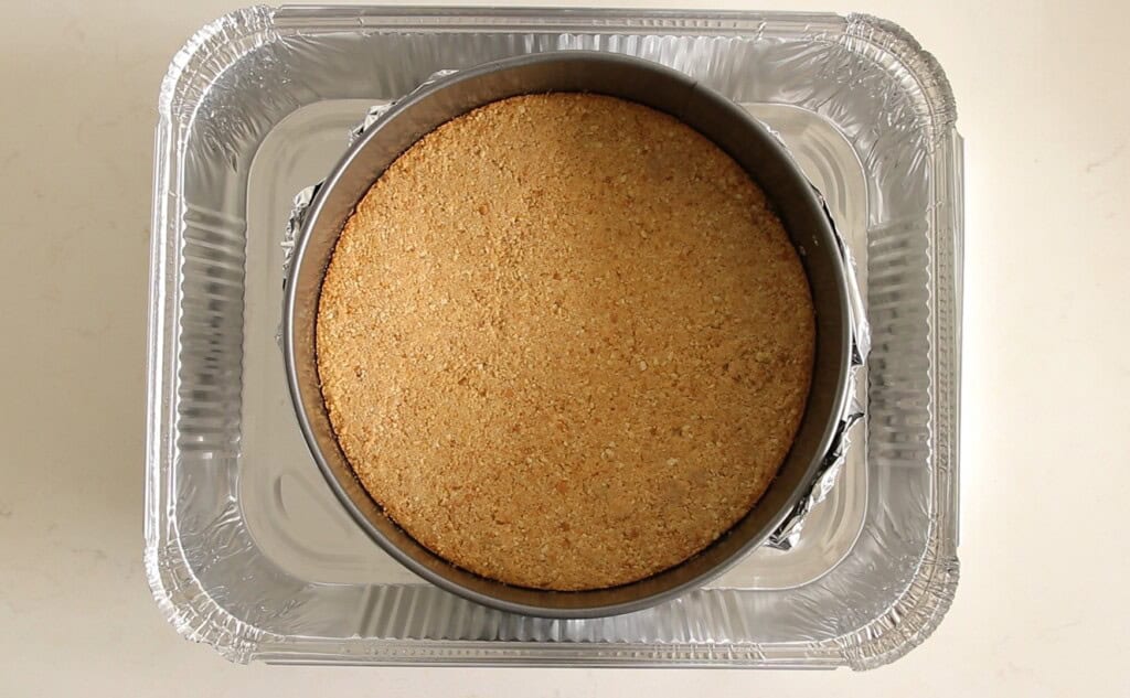 A round pie crust in a springform pan sits inside a larger disposable aluminum tray, viewed from above on a light surface.