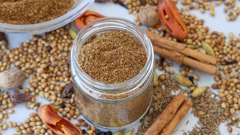 A jar of ground spice blend is surrounded by whole spices, including cinnamon sticks, cardamom pods, coriander seeds, nutmeg, cumin seeds, and mace, on a white surface.
