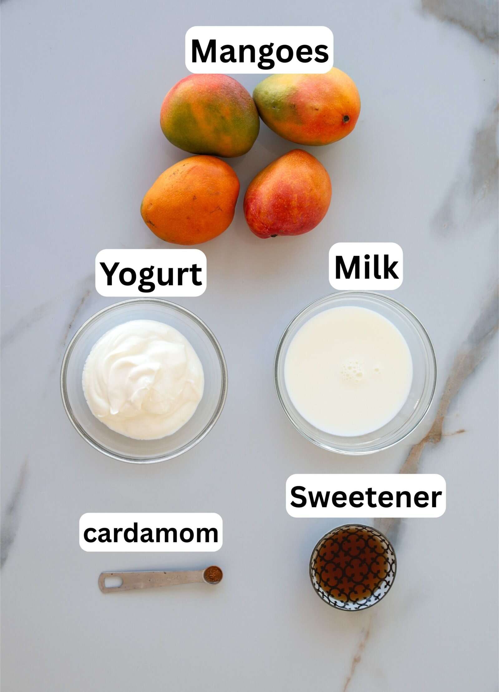 Overhead view of ingredients for mango lassi: three whole mangoes, a bowl of yogurt, a bowl of milk, a small container of sweetener, and a measuring spoon with cardamom, all labeled.