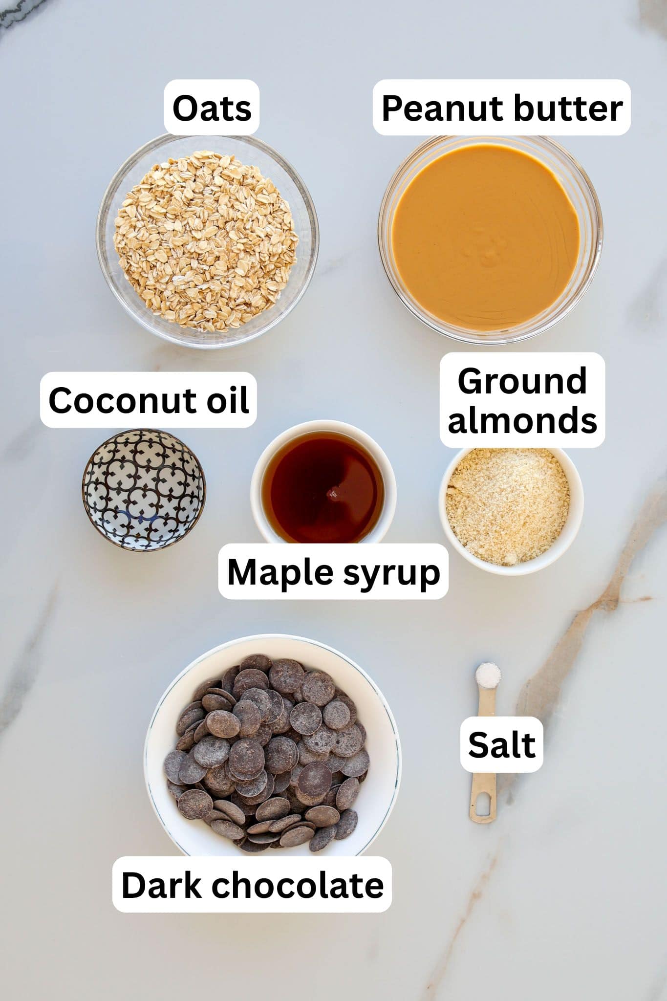 Seven labeled bowls and a measuring spoon on a white surface, each containing: oats, peanut butter, coconut oil, maple syrup, ground almonds, dark chocolate, and salt.