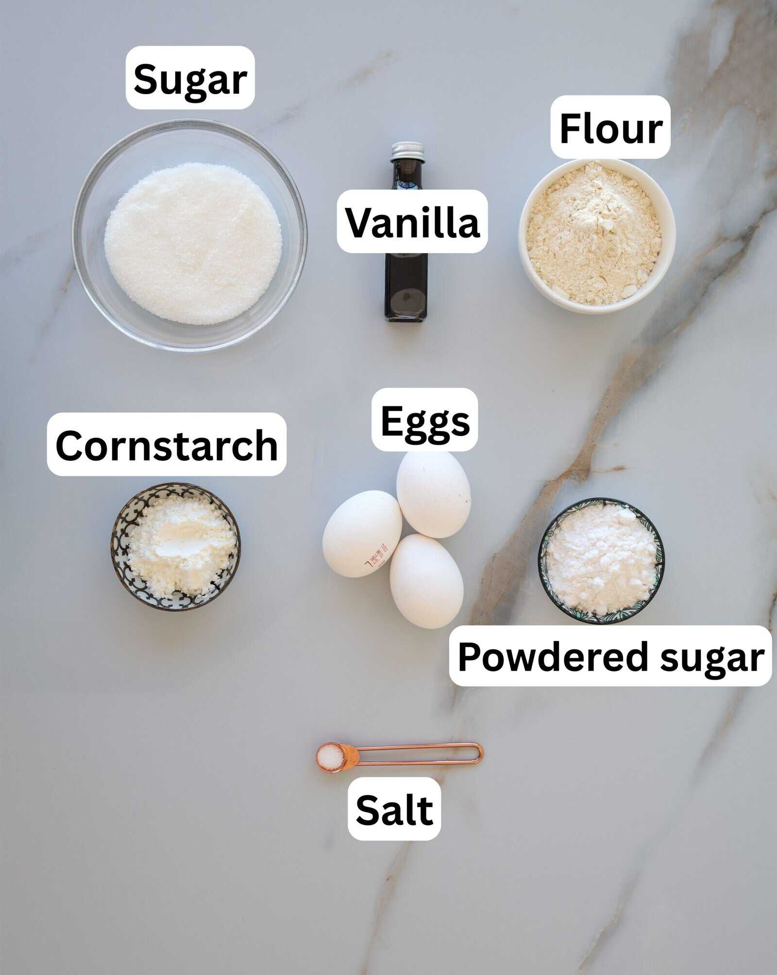 A flat lay of baking ingredients labeled: sugar, vanilla, flour, cornstarch, eggs, powdered sugar, and salt on a light marble surface.