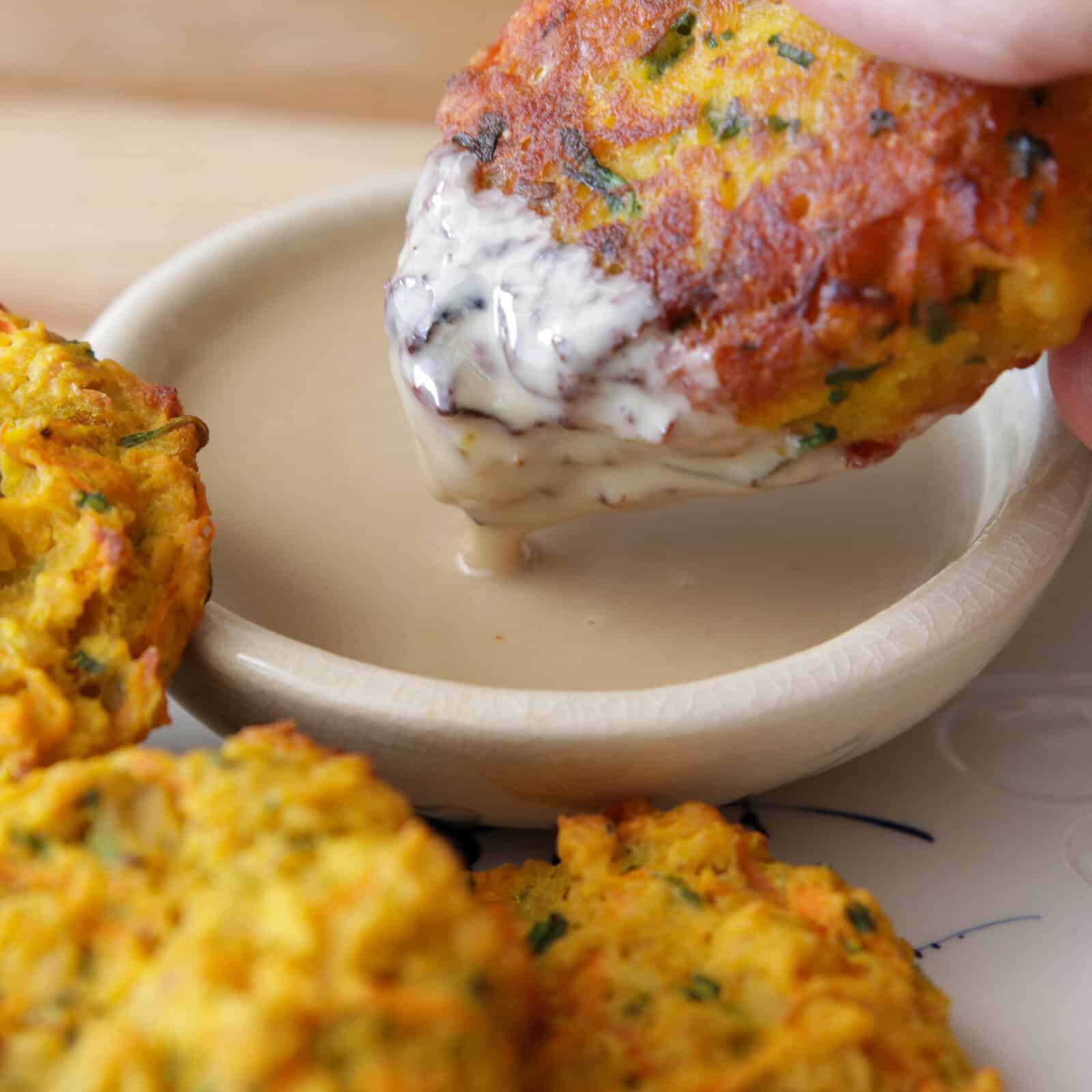 A hand dips a golden, crispy vegetable fritter into a creamy, light-colored tahini sauce in a small bowl, with more fritters visible on the plate nearby.
