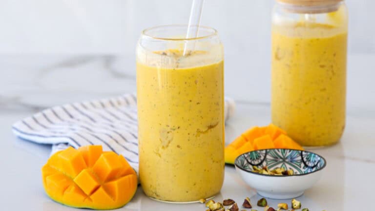 A glass of mango lassi with a straw sits on a white surface, surrounded by sliced fresh mango, chopped pistachios in a small bowl, and a second jar of lassi in the background. A striped cloth is also nearby.