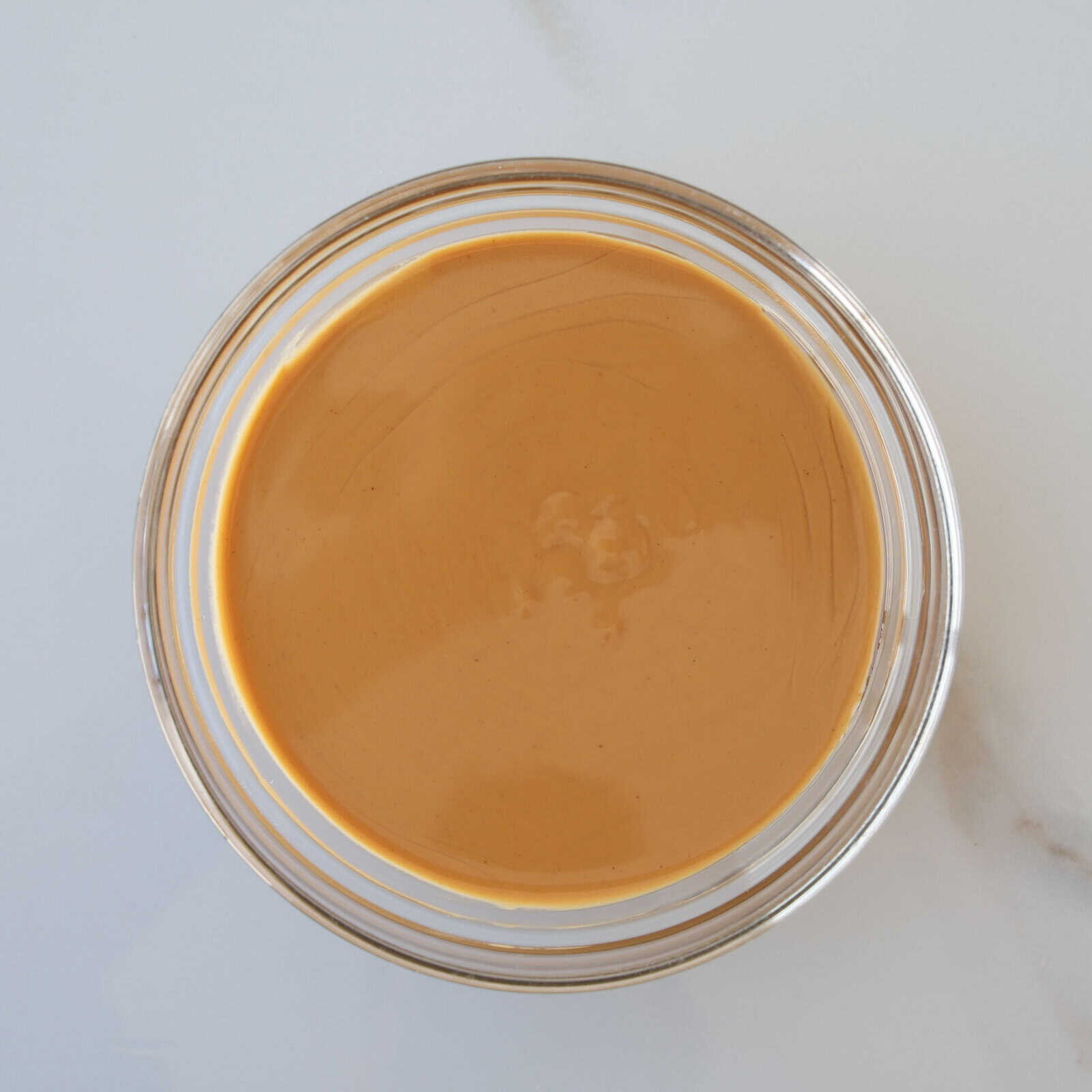 A clear glass bowl filled with smooth, creamy peanut butter sits on a white surface.