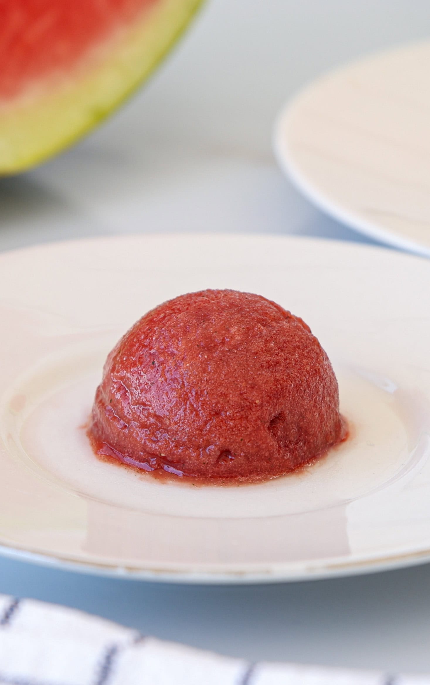 A single scoop of red watermelon sorbet sits on a white plate, with a blurred slice of watermelon and another white plate in the background.