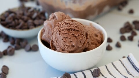 A white bowl filled with scoops of homemade chocolate ice cream sits on a white surface, surrounded by scattered chocolate chips and a striped cloth. A bowl of chocolate chips and a container of ice cream are in the background.