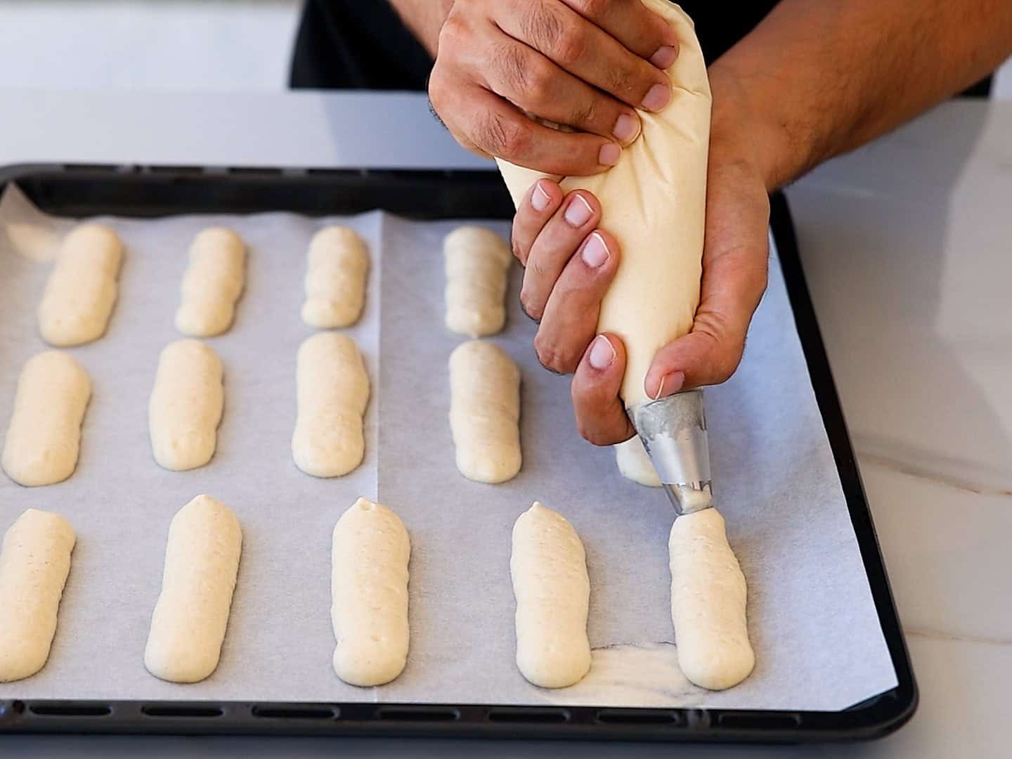 A person uses a piping bag to pipe long strips of dough onto a parchment-lined baking tray, preparing pastry or cookies.