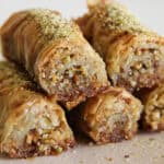 Close-up of several pieces of rolled baklava, filled with chopped nuts and topped with ground pistachios, stacked on a light surface. The pastry looks crispy and golden brown.