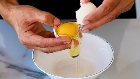 Close-up of hands cracking an egg over a white bowl, with the yolk and egg white falling into the bowl on a marble countertop.