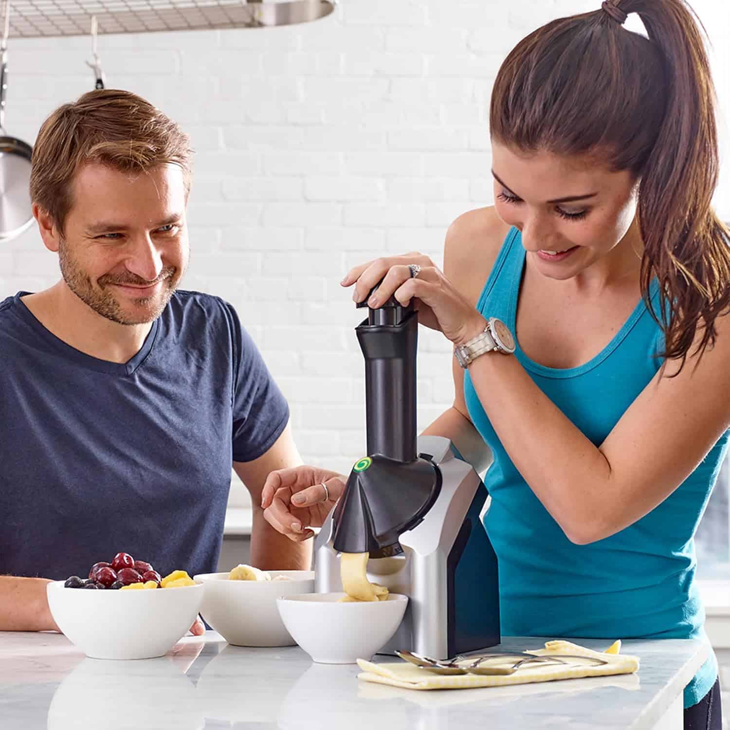 A woman uses a kitchen appliance to make banana dessert while a man watches and smiles. Bowls of fruit, including bananas and grapes, sit on the counter in a bright kitchen.