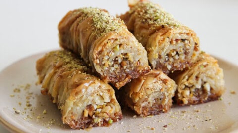 Easy Baklava Rolls Recipe – Crispy Greek Saragli with Three Nuts Six pieces of rolled baklava filled with chopped nuts are stacked on a plate, with golden brown, flaky layers and a sprinkle of ground pistachios on top.
