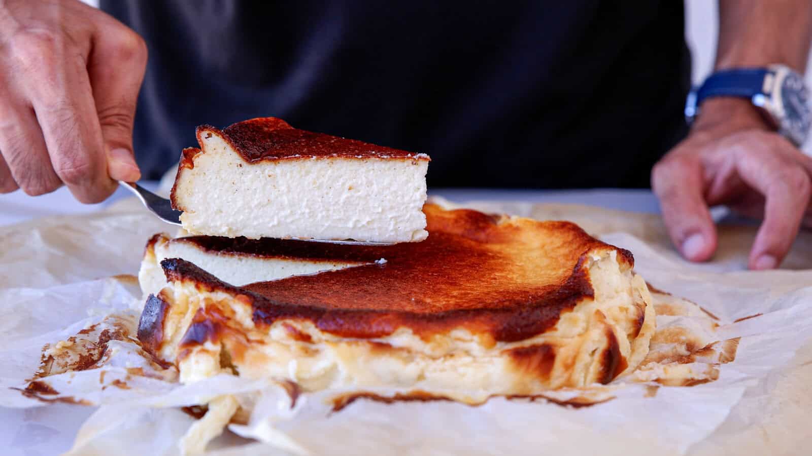 A person lifts a slice of creamy, burnt cheesecake from a parchment-lined whole cake, revealing its caramelized top and soft interior.