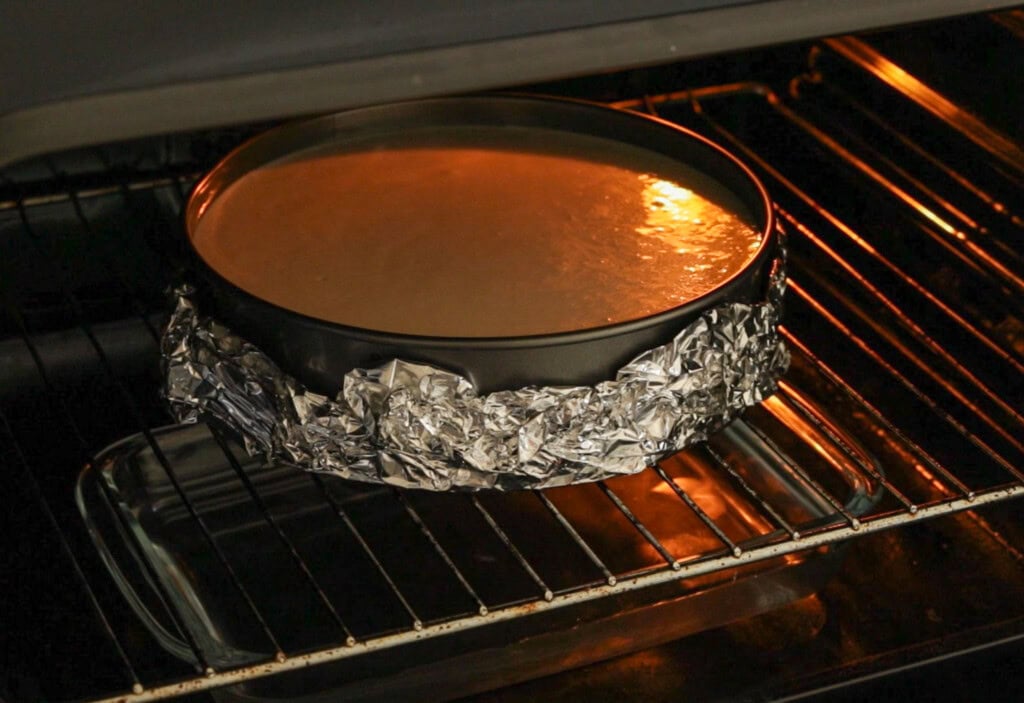 A round springform pan wrapped in aluminum foil sits on an oven rack, baking a cheesecake. The pan is placed inside a glass dish, likely for a water bath. The cheesecake batter appears smooth and creamy.