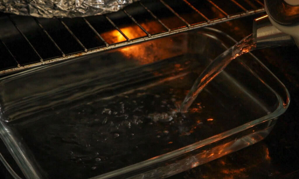 A close-up of water being poured from a kettle into a glass baking dish placed on the rack inside an oven, with a warm orange glow reflecting off the glass.