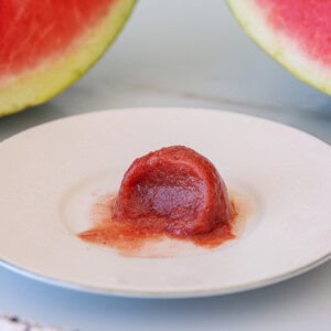 A small, round scoop of red watermelon sorbet sits on a white plate, with two slices of fresh watermelon in the background.