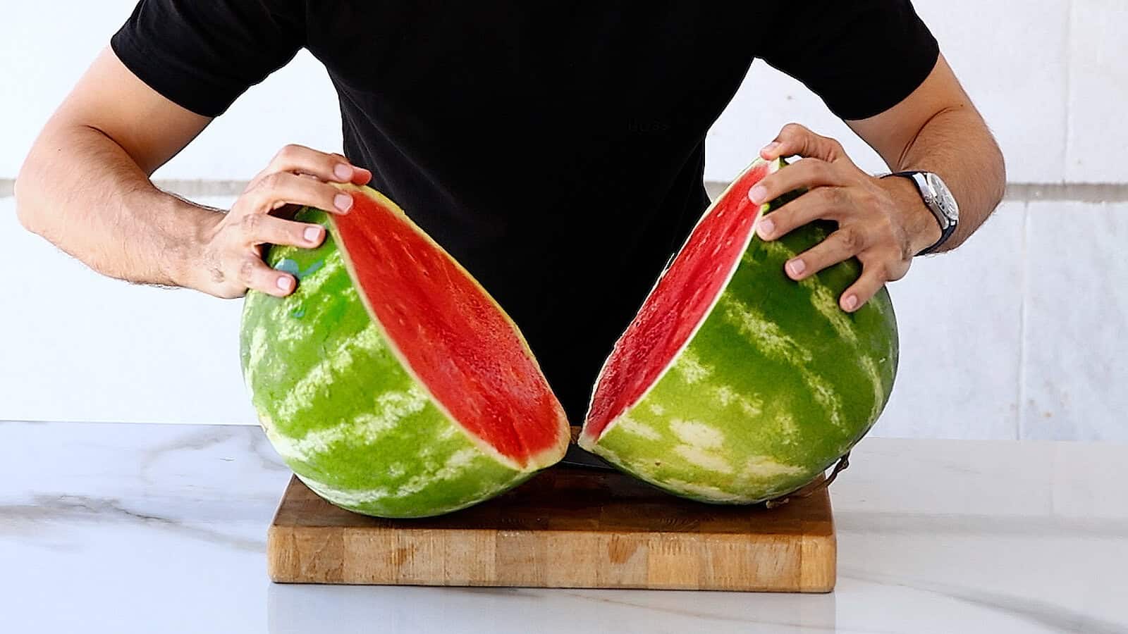 A person in a black shirt splits a watermelon in half on a wooden cutting board, revealing the bright red fruit inside.
