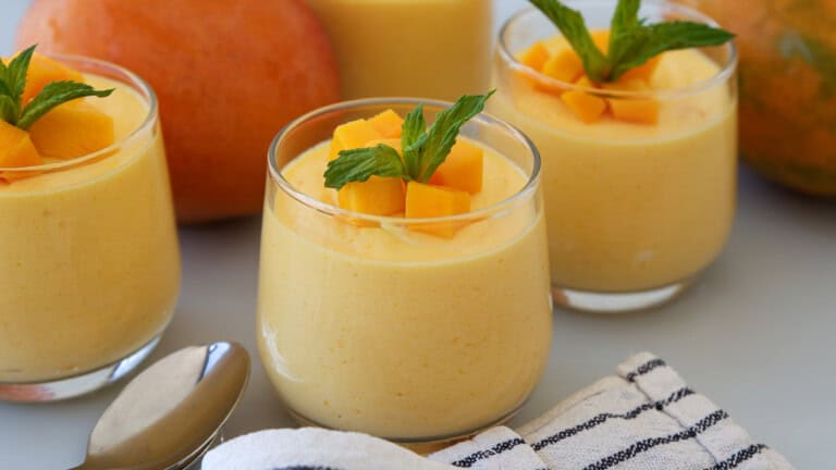 Three glasses of creamy mango mousse topped with fresh mango cubes and mint leaves, placed beside a spoon, a striped napkin, and whole mangoes in the background.