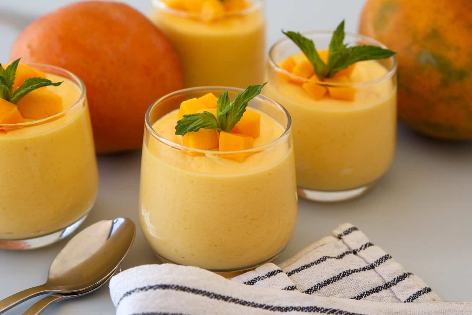Four glass cups filled with creamy mango mousse, topped with diced mango and mint leaves. A striped napkin and two spoons are in the foreground, with whole mangoes in the background.