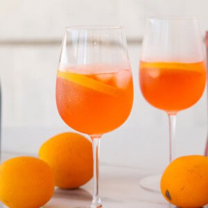 Two wine glasses filled with an orange cocktail, ice cubes, and orange slices, with whole oranges placed on a white surface in the foreground and background.