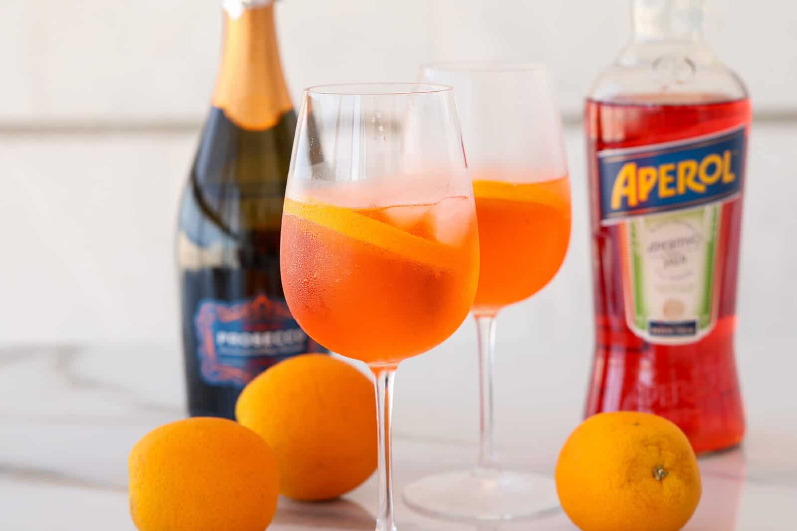 Two glasses of orange Aperol spritz with orange slices and ice, surrounded by whole oranges, a bottle of Prosecco, and a bottle of Aperol on a white surface.