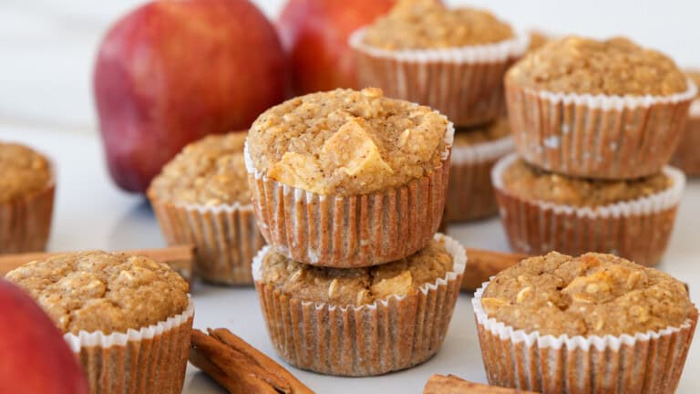 A close-up of apple oatmeal muffins stacked and arranged with apples and cinnamon sticks in the background, displayed on a light surface.
