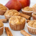 Apple muffins in paper liners are arranged on a white surface with cinnamon sticks nearby and apples in the background, suggesting a spiced apple flavor.