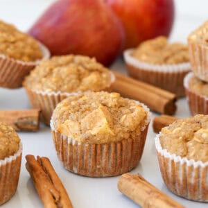 Apple muffins in paper liners are arranged on a white surface with cinnamon sticks nearby and apples in the background, suggesting a spiced apple flavor.
