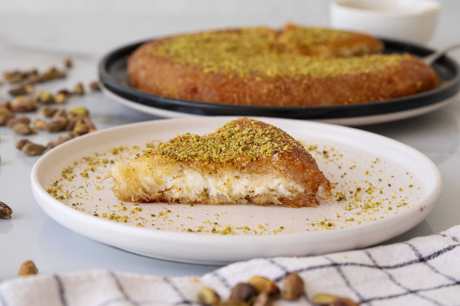 A slice of knafeh topped with ground pistachios sits on a white plate, with the remaining dessert and whole pistachios in the background. A checkered cloth is partially visible in the foreground.