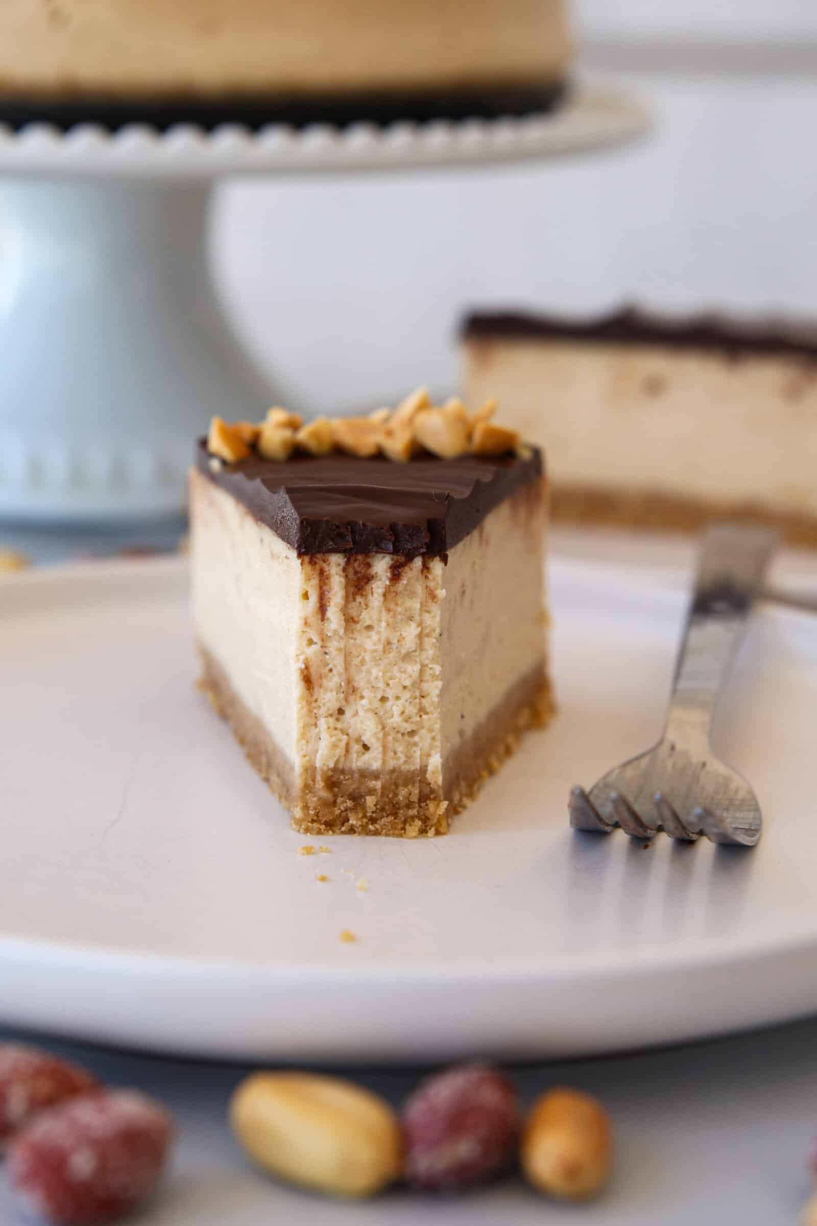 A slice of a peanut butter cheesecake with a chocolate topping and sprinkled peanuts sits on a white plate, with a fork nearby and whole peanuts and strawberries in the foreground. A whole cheesecake is in the background on a cake stand.